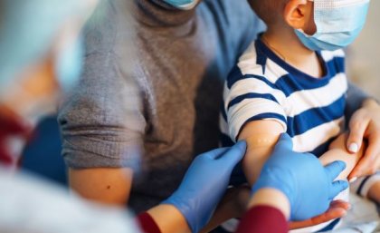 Gloved hands are smoothing a bandaid onto a child's upper arm. The child is on the lap of an adult carer.